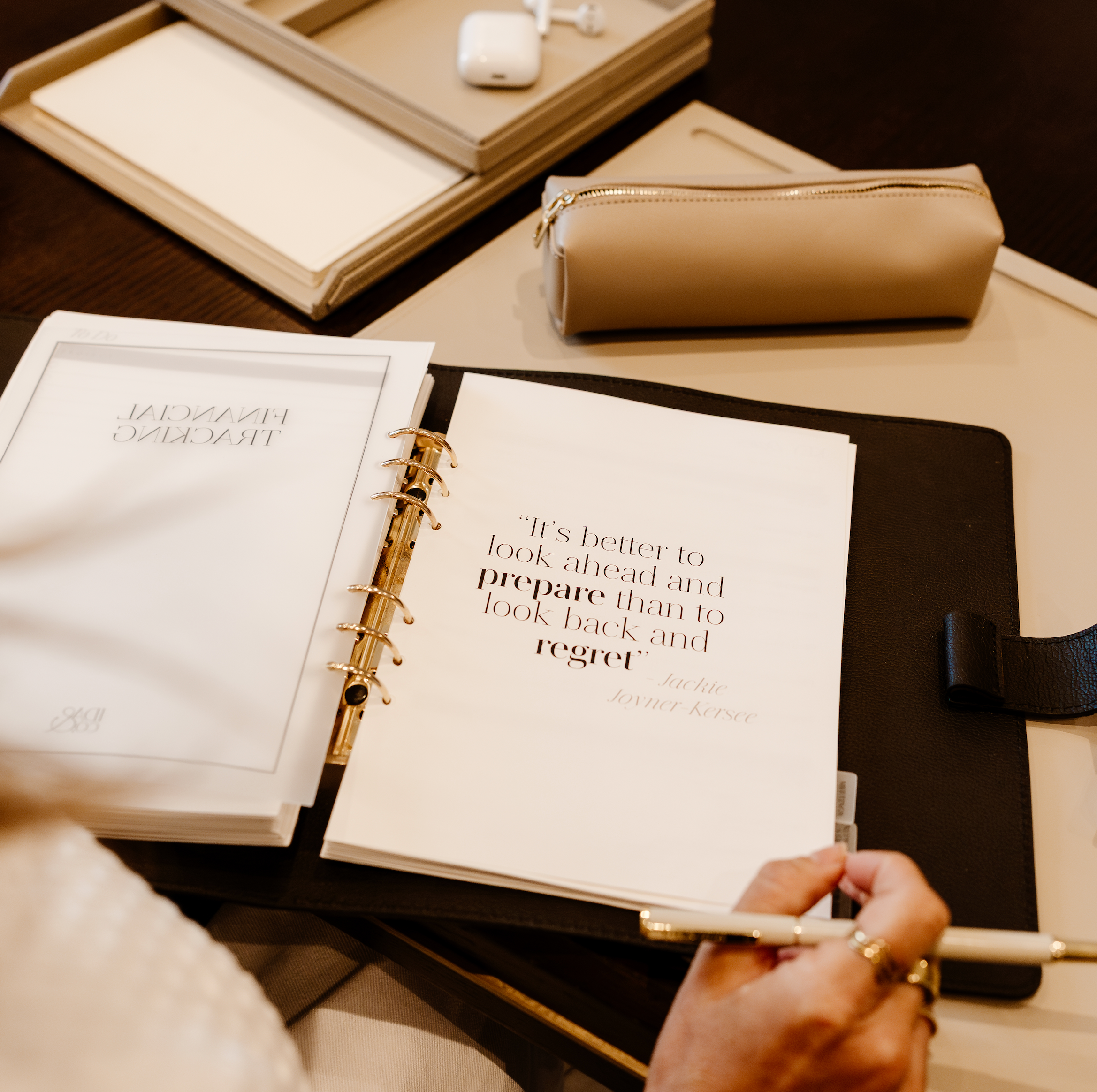 Person holding a pen over an open planner with a quote, on a desk with books and a notebook.