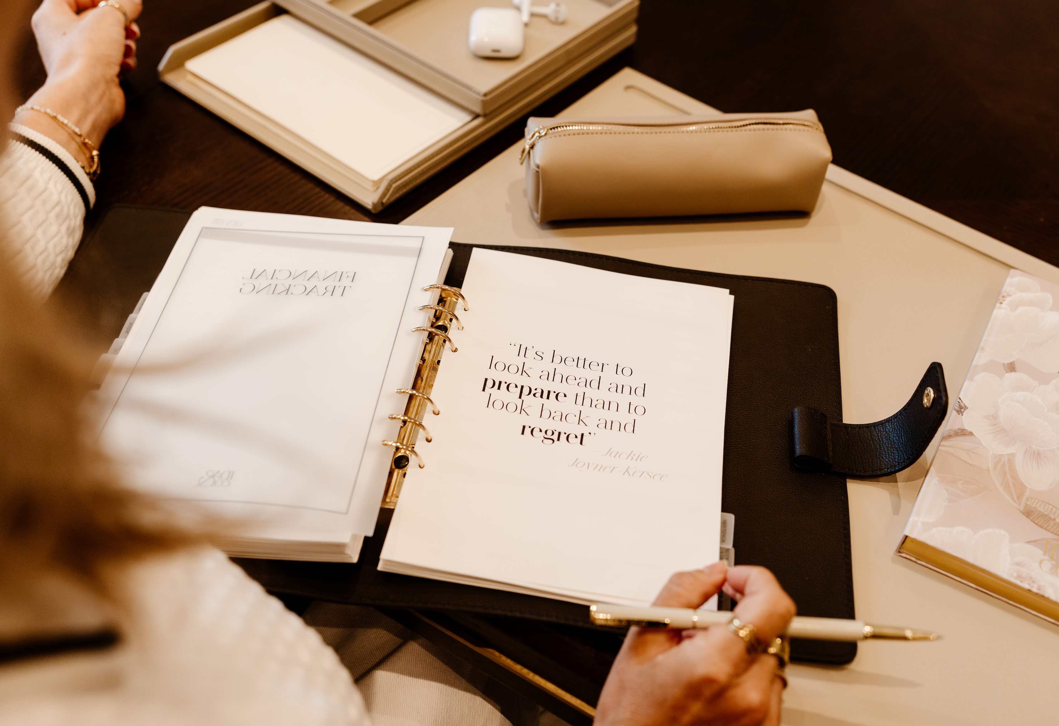 Person holding a pen over an open planner with a quote, on a desk with books and a notebook.