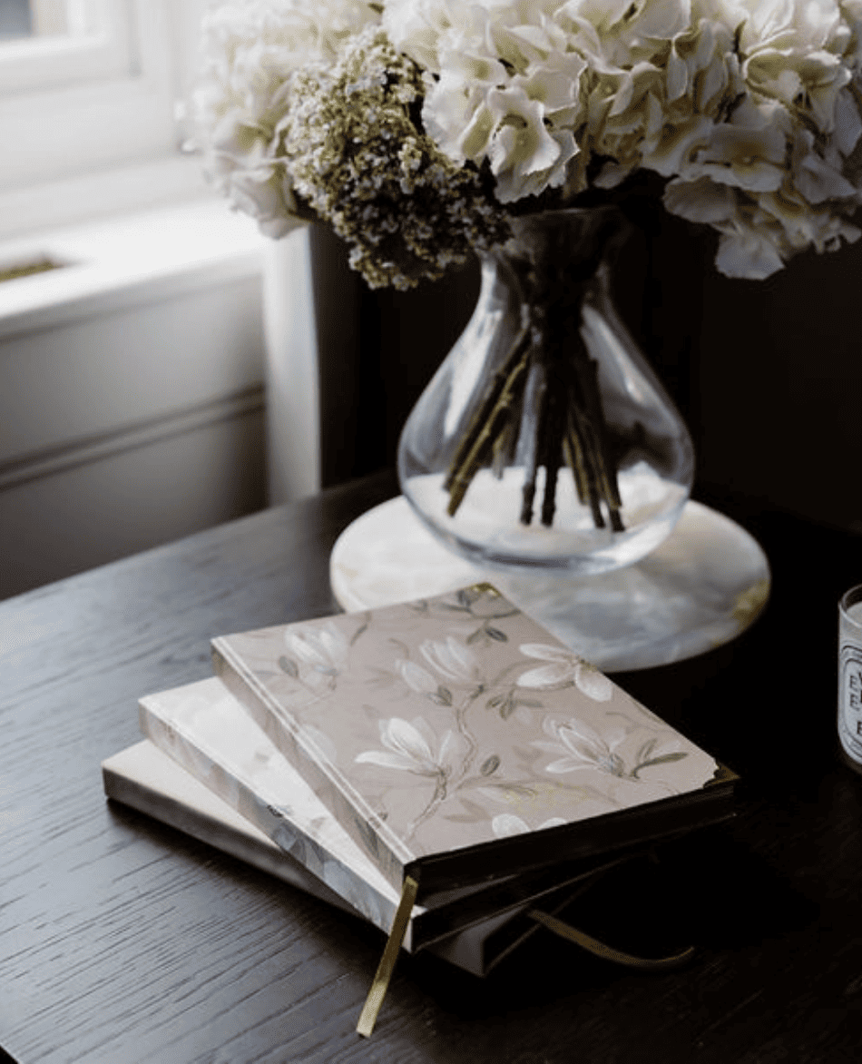 Two floral-patterned books on a wooden surface with a vase of white flowers in the background.