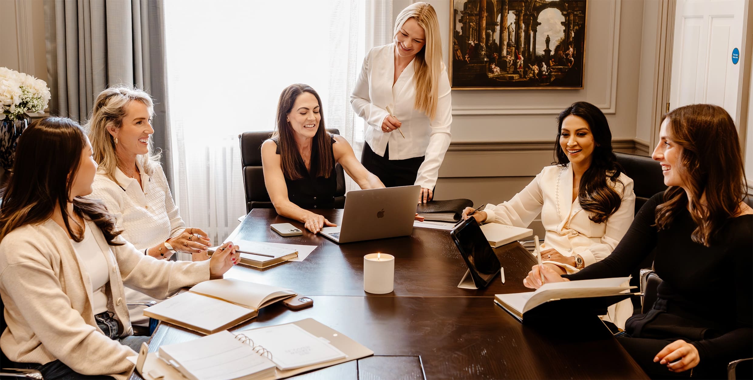 Group of women in a meeting around a conference table with a woman standing and speaking.